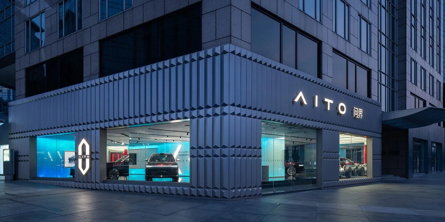 AITO showroom at dusk with a textured grey facade, illuminated AITO sign and cars visible through the glass
